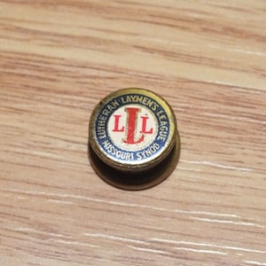 May include: A gold-coloured pin with a blue circle and white lettering that reads "Lutheran Laymens League Missouri Synod". The letters "LL" are in red and are centred in the circle.