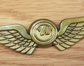 Puede incluir: Pin de metal de color dorado con una "W" estilizada en un círculo y alas a cada lado.