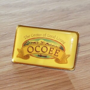 Può includere: Uno spillo in metallo dorato con sfondo giallo e il testo "The Center of Good Living" e "OCOEE" in uno stendardo.