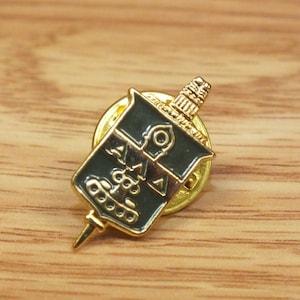 May include: Gold-coloured pin with a black and green shield-shaped design. The shield features the letters "AMA" and a stylised image of a building. The pin has a pointed tip.