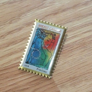 May include: A gold-colored US postage stamp with a colorful illustration of a woman and the text "Breast Cancer" and "Fund the Fight". The stamp also says "USA First-Class".