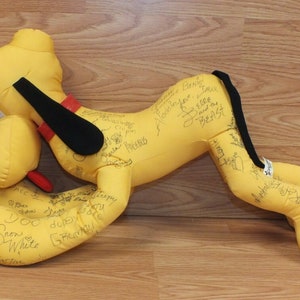 Peut inclure: Un chien en peluche jaune avec des oreilles noires et une queue noire. Le chien est couché sur le côté, la tête tournée vers la gauche. Le chien est couvert de signatures, dont "Snow White" et "The Beast".