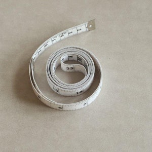 May include: A white measuring tape rolled up in a spiral on a beige surface. The tape has black markings with numbers from 1 to 33.