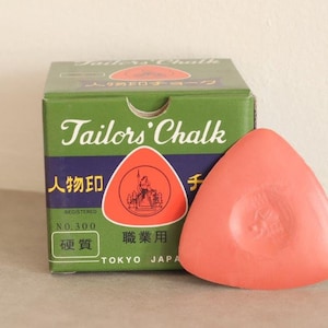 May include: A green box of tailor's chalk with the text "Tailor's Chalk" and "Registered No. 300" in white. The box also has Japanese text. A pink triangular piece of tailor's chalk is in front of the box.
