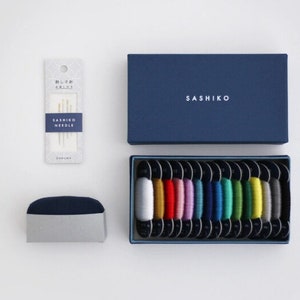 May include: A blue box with the word "SASHIKO" printed on it, containing a set of 16 spools of colourful embroidery floss. A smaller blue box with a white lid contains a set of needles. A separate package contains a set of needles labelled "SASHIKO NEEDLE".