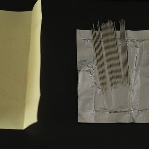 May include: A package of silver sewing needles wrapped in a silver foil packet. The needles are arranged in a neat row and are ready to be used for sewing projects.