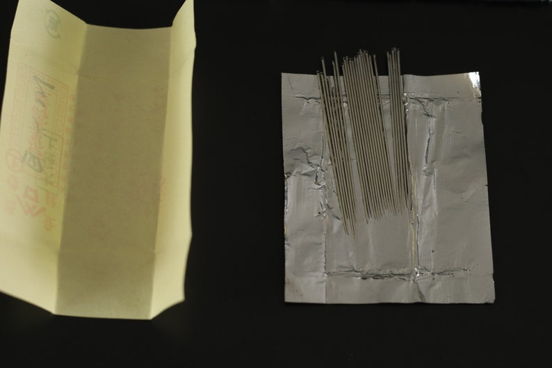 May include: A package of silver sewing needles wrapped in a silver foil packet. The needles are arranged in a neat row and are ready to be used for sewing projects.
