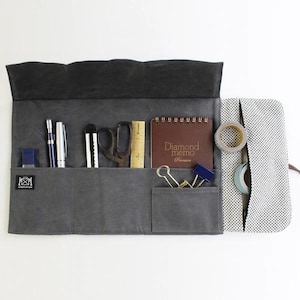 May include: A gray canvas roll-up pouch with multiple pockets and compartments. The pouch is open and contains a variety of stationery items, including pens, pencils, scissors, a ruler, a small notebook, tape, and paper clips. The notebook has the text "Diamond memo" on the cover.