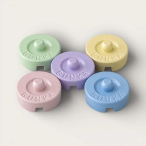 May include: Five circular pastel-colored objects with the word BUNKA embossed on the top surface. Each piece features a central raised knob and a small rectangular notch at the base. The colors include light green, yellow, lavender, pink, and blue.