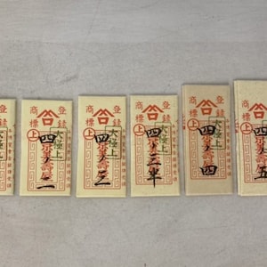May include: Six small, rectangular, cream-colored paper tags with red borders and black text. Each tag has the text "商標登録" in Japanese, which translates to "Registered Trademark" in English. The tags also have a red square with a white circle inside, and the text "合" in black. The text below the red square varies on each tag, but all include the text "壽屋".
