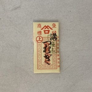 Peut inclure: Un petit paquet de papier rectangulaire de couleur crème avec du texte rouge et noir. Le texte indique "商合", "精", "上", et "真宗", avec un sceau rouge qui dit "合" en blanc.