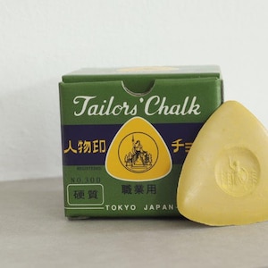 May include: A green box of tailor's chalk with the text "Tailor's Chalk" in white lettering. The box also has Japanese text and the words "Registered No. 300" and "Tokyo Japan". A yellow triangular piece of tailor's chalk is next to the box.