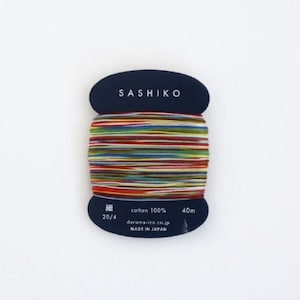 May include: A spool of colorful Sashiko thread, labeled "Sashiko" and "cotton 100%". The thread is wound around a blue card with the text "20/4", "daruma-ito.co.jp", and "MADE IN JAPAN".