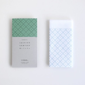 Peut inclure: Un livre vert et blanc avec le texte "Daruma Sashiko Sampler" et "十文字刺し" sur la couverture. À côté du livre se trouve un tissu blanc avec un motif géométrique bleu et blanc.