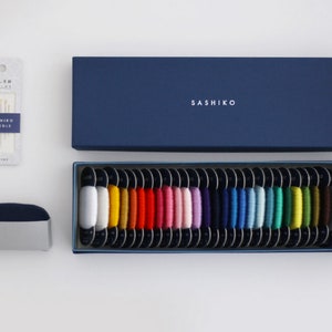 Pode incluir: Uma caixa azul com a palavra "SASHIKO" impressa na tampa. A caixa está aberta e contém 36 carretéis de fio de várias cores. Os carretéis estão dispostos em filas e são etiquetados com números. Também há um pequeno pacote de agulhas e um dedal.