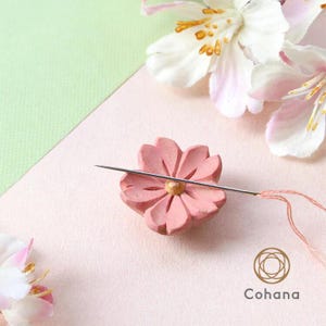 Può includere: Un portaghiaccio in legno rosa a forma di fiore con ago e filo. L'ago è inserito al centro del fiore. Lo sfondo è rosa tenue e verde con fiori bianchi e rosa.