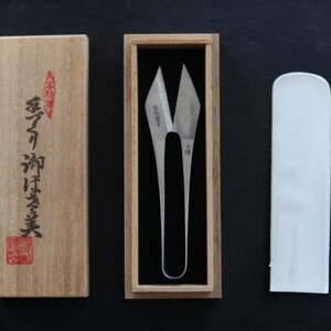 May include: A pair of silver-coloured fabric shears in a wooden box with a Japanese inscription. The shears are in a white fabric sheath.