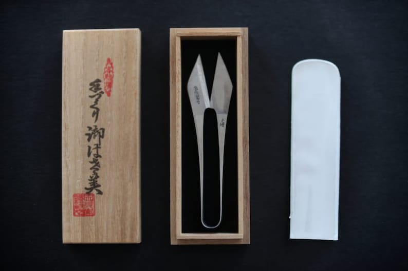 May include: A pair of silver-coloured fabric shears in a wooden box with a Japanese inscription. The shears are in a white fabric sheath.