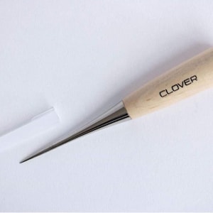 May include: A Clover awl with a wooden handle and a silver metal tip. The handle is light-coloured with the brand name printed in black. A white plastic cap is next to the awl. This tool is used for sewing and crafting.