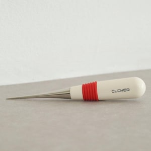Könnte beinhalten: Ein weißes und rotes Nähwerkzeug mit einer spitzen Spitze und einem roten Gummibeschlag. Das Werkzeug ist auf dem Griff mit "CLOVER" beschriftet.