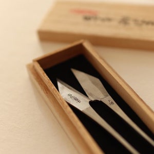May include: A pair of silver-coloured shears with black handles are displayed in a wooden box. The shears have Japanese text on the blades.
