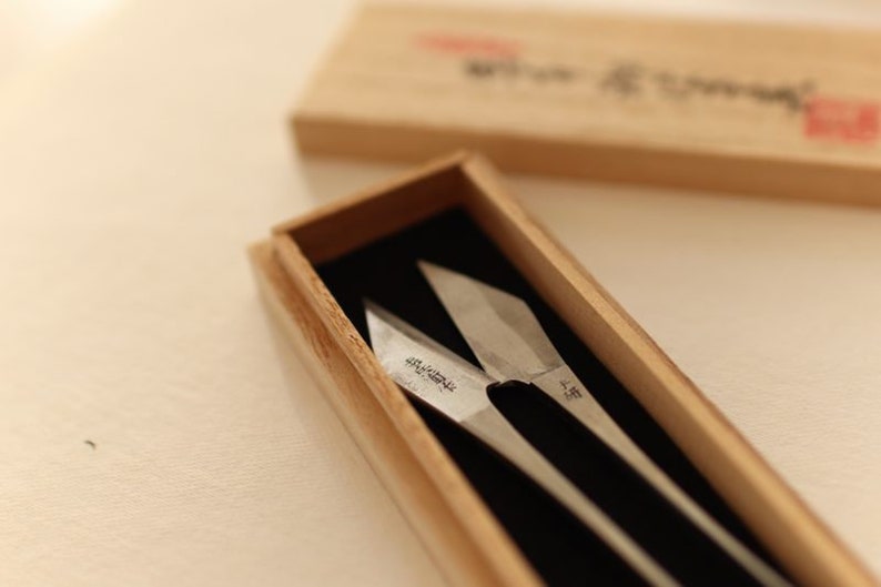 May include: A pair of silver-coloured shears with black handles are displayed in a wooden box. The shears have Japanese text on the blades.