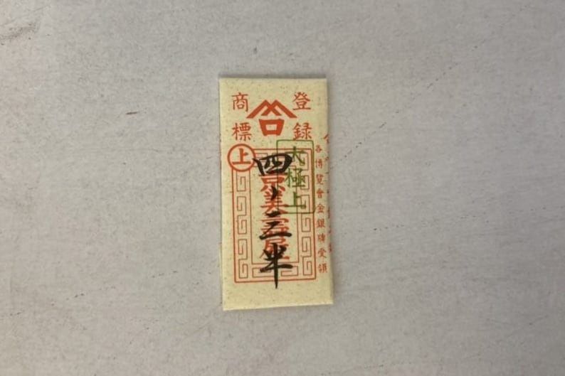 May include: A small, rectangular, cream-colored paper packet with red Chinese characters that read "Shang Hai Jin", which translates to "Shanghai Gold". The packet has a red border and a gold seal with a white triangle inside.