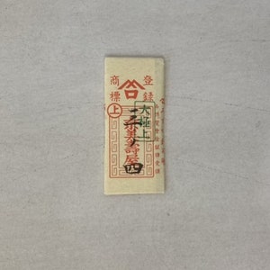 May include: A small, rectangular, brown paper packet with red and green Chinese characters printed on it. The text reads "商  合  金  銀  上  本  業  屋".