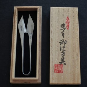 May include: A pair of silver-coloured, pointed scissors in a wooden box with Japanese writing on the lid. The writing on the lid reads "香蘭  和鋏 実業".