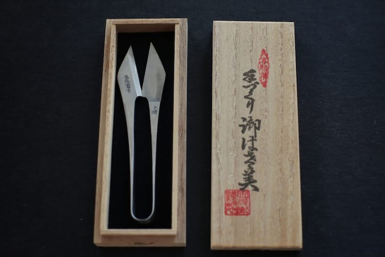 May include: A pair of silver-coloured, pointed scissors in a wooden box with Japanese writing on the lid. The writing on the lid reads "香蘭  和鋏 実業".