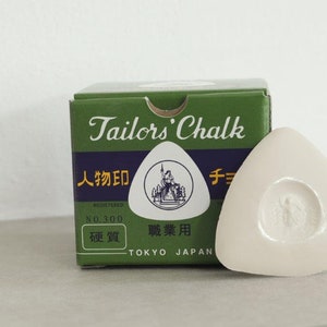 May include: A green box with the text "Tailors' Chalk" in white lettering. The box also has Japanese text and the words "Registered No. 300" and "Hard Quality". A white triangular piece of tailor's chalk is next to the box.