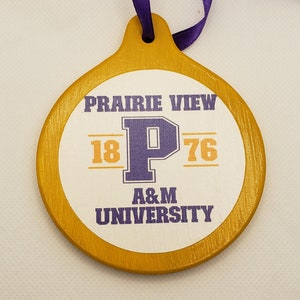 May include: A round, wooden ornament painted gold with a white circle in the centre. The circle has the text "Prairie View 18 P 76 A&M University" in purple and orange lettering.