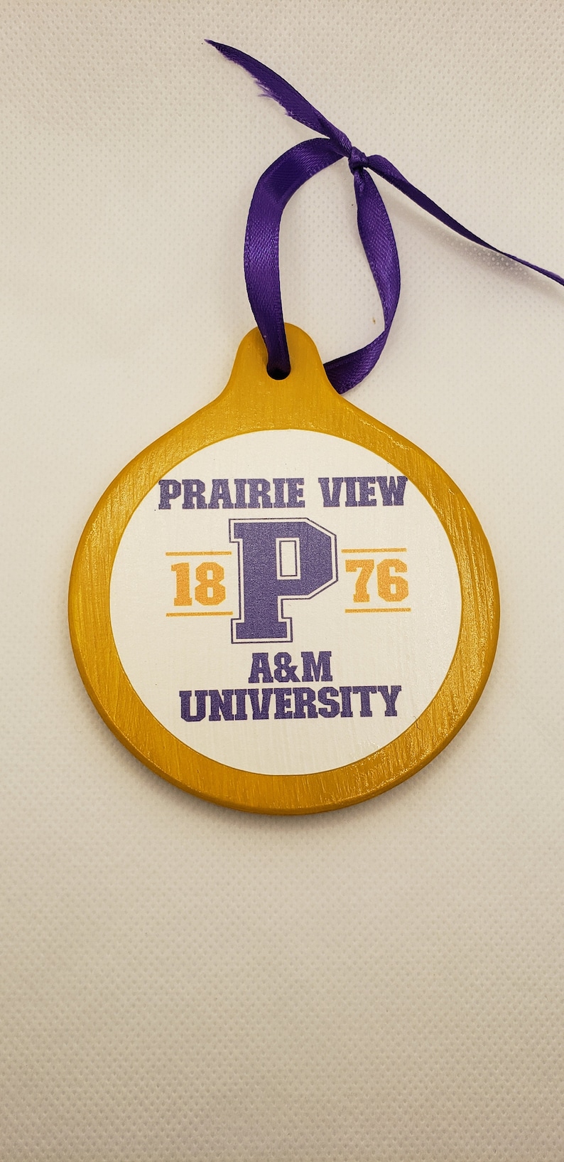 May include: A round, wooden ornament painted gold with a white circle in the centre. The circle has the text "Prairie View 18 P 76 A&M University" in purple and orange lettering.