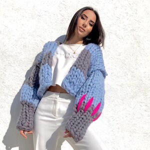 May include: A chunky knit cardigan in shades of blue and gray, with bright pink accents on the sleeves. The oversized sweater is paired with a white crop top and white pants. The cardigan's thick yarn creates a textured, cozy look.
