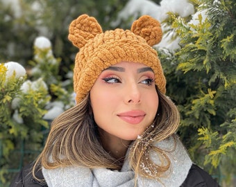 May include: A woman wearing a brown fuzzy knit hat with bear ears, a gray scarf, and a black puffer jacket. She is standing in front of a snowy evergreen tree.