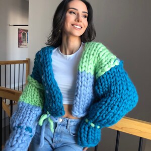 May include: A chunky knit cardigan in shades of blue and green. The oversized sweater features a cropped design with long sleeves and a tie closure. The color-blocked pattern includes teal, light blue, and lime green sections, creating a cozy and stylish look.