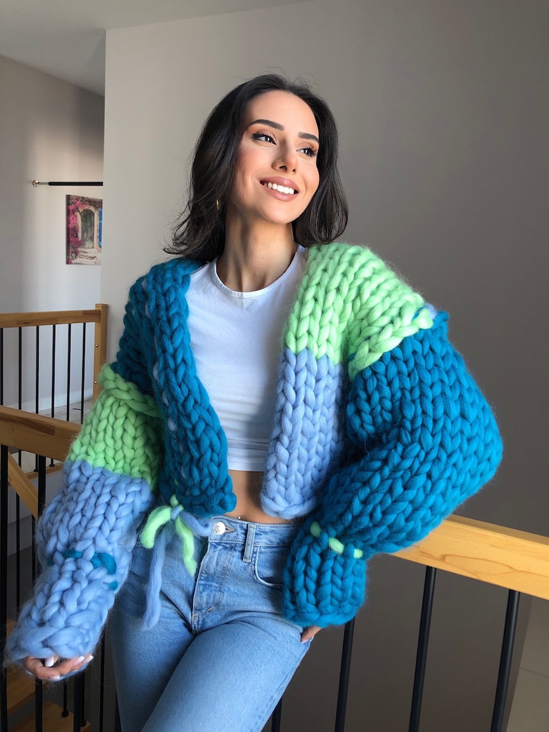 May include: A chunky knit cardigan in shades of blue and green. The oversized sweater features a cropped design with long sleeves and a tie closure. The color-blocked pattern includes teal, light blue, and lime green sections, creating a cozy and stylish look.