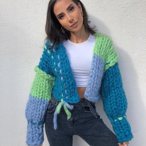 May include: A chunky knit cardigan in shades of blue and green. The oversized sweater features a drawstring closure and a cropped design. The sleeves are long and voluminous, and the knit has a thick, textured appearance.