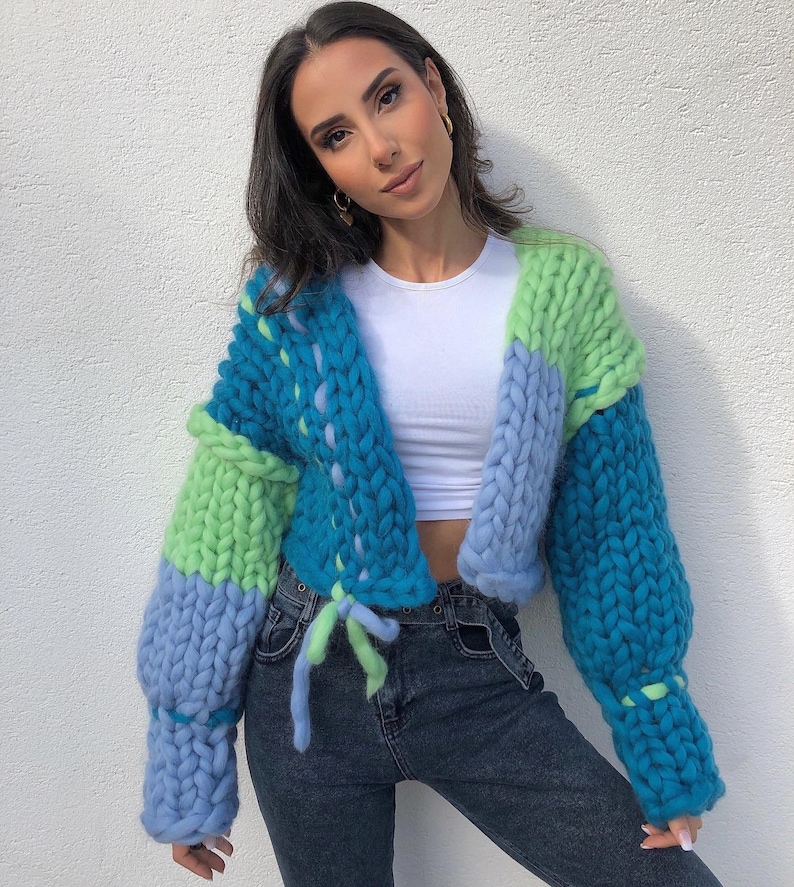 May include: A chunky knit cardigan in shades of blue and green. The oversized sweater features a drawstring closure and a cropped design. The sleeves are long and voluminous, and the knit has a thick, textured appearance.