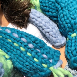 May include: Close-up of chunky knit textiles in shades of teal, light blue, and green. The knitted pieces have a thick, textured appearance, with visible stitches and varying color patterns. The textiles are arranged in a layered composition.