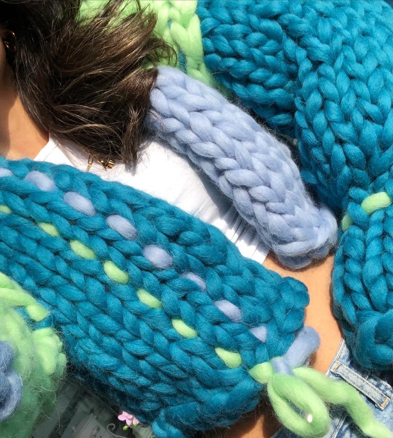May include: Close-up of chunky knit textiles in shades of teal, light blue, and green. The knitted pieces have a thick, textured appearance, with visible stitches and varying color patterns. The textiles are arranged in a layered composition.