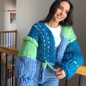 May include: A chunky knit cardigan in shades of blue and green. The cropped sweater features a tie closure and oversized sleeves. The knitwear is made of thick yarn, creating a textured appearance.