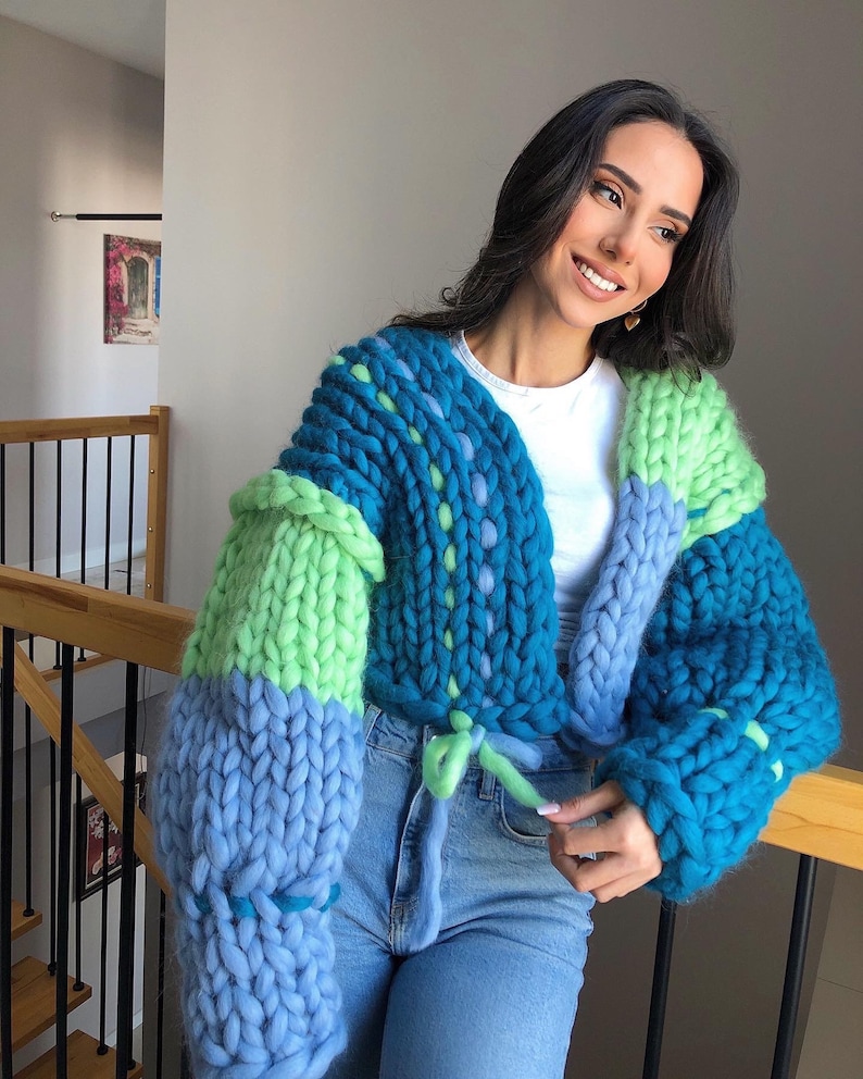 May include: A chunky knit cardigan in shades of blue and green. The cropped sweater features a tie closure and oversized sleeves. The knitwear is made of thick yarn, creating a textured appearance.