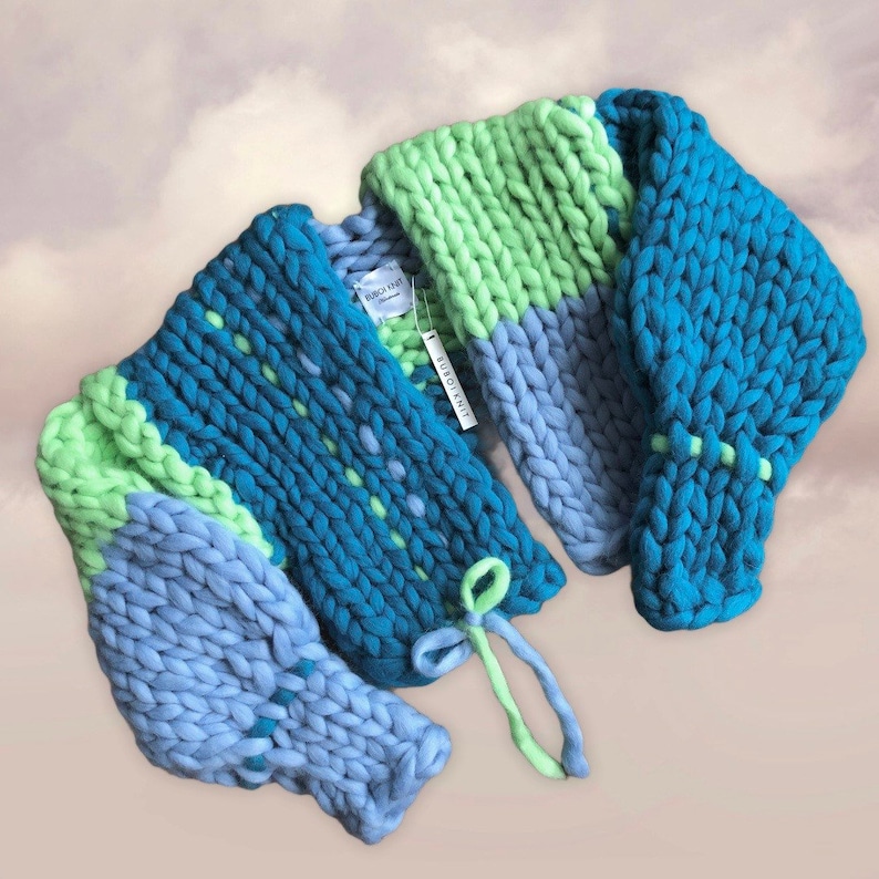 May include: A chunky knit sweater in shades of blue and green. The sweater features a color-blocked design with sections of light blue, teal, and lime green. The sleeves are thick and the sweater has a tie closure. The tag reads "SUBOIKNIT".