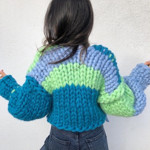 May include: A chunky knit sweater in shades of teal, light blue, and green. The sweater features a color-blocked design with a ribbed hem and oversized sleeves. The back view showcases the textured, hand-knitted construction.