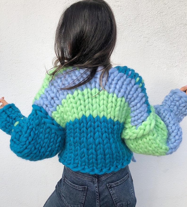 May include: A chunky knit sweater in shades of teal, light blue, and green. The sweater features a color-blocked design with a ribbed hem and oversized sleeves. The back view showcases the textured, hand-knitted construction.
