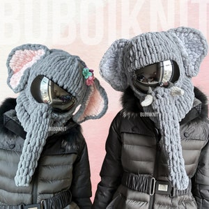 May include: Two gray knitted elephant hats with pink ears and a gray knitted elephant trunk. The hats have a black ski goggle attached to the front. The hats are worn by two people. The text "Bubooknit" is on the hats.