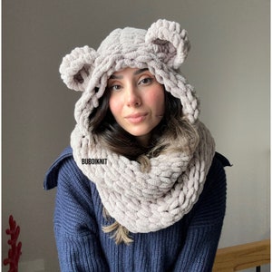 May include: A light pink hooded scarf with bear ears. The scarf is made of a soft, fluffy yarn and is perfect for keeping warm in the winter. The scarf is worn by a person with long brown hair. The text "BUBOIKNIT" is visible on the scarf.