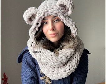 May include: A light pink hooded scarf with bear ears. The scarf is made of a soft, fluffy yarn and is perfect for keeping warm in the winter. The scarf is worn by a person with long brown hair. The text "BUBOIKNIT" is visible on the scarf.