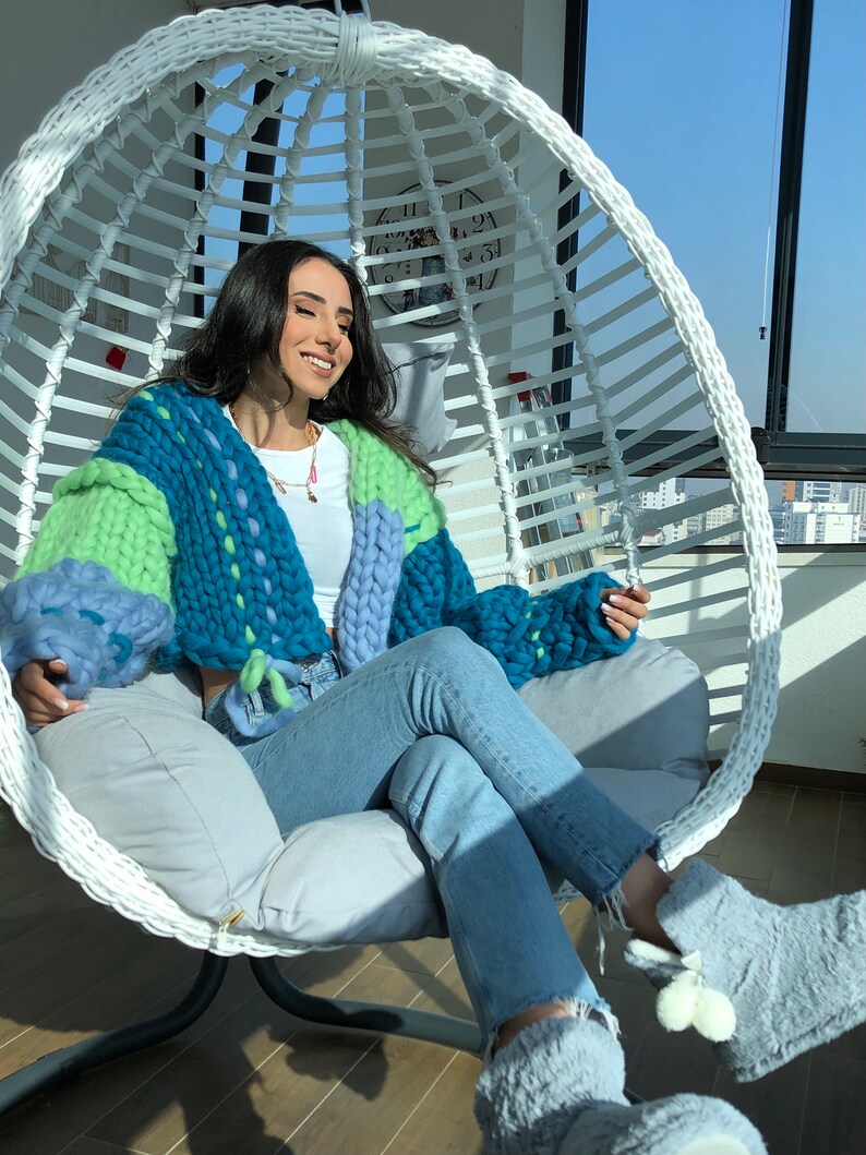 May include: A chunky knit cardigan in shades of blue and green, worn with light wash jeans and gray slippers. The oversized sweater features a patchwork design and is made of thick yarn. The person is seated in a white hanging chair.
